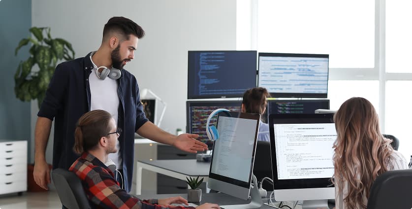 Developers collaborating at computer workstations in a modern office, reviewing JavaScript code on large monitors and demonstrating how a team uses JavaScript and JSON in a Django web project for web development.