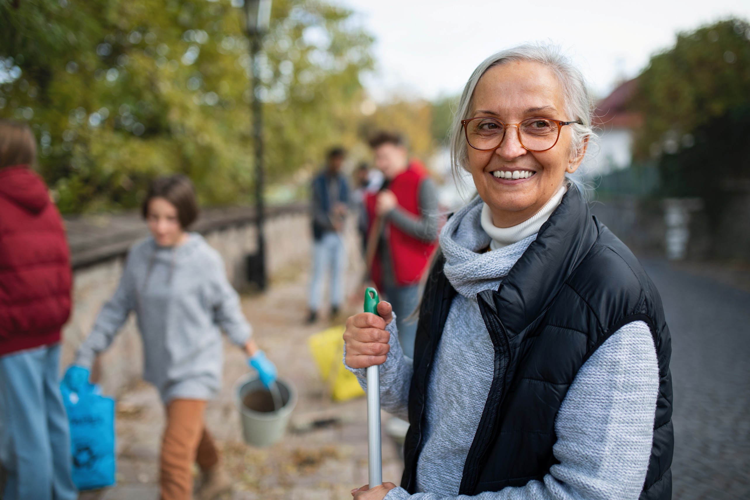 A smiling elderly woman holding a broom with volunteers cleaning a public space in the background