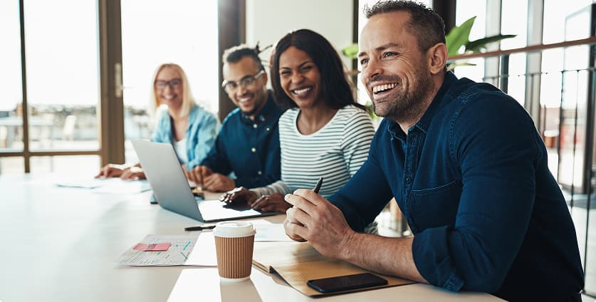 Gruppe von Kolleginnen und Kollegen, die lächeln und an einer Besprechung in einem hellen, modernen Büro teilnehmen.